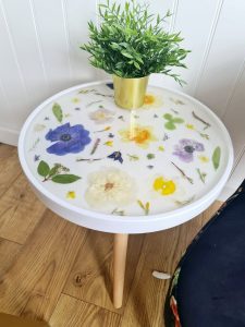 Round white resin side table with real dried flowers and leaves embedded in a glossy epoxy surface, featuring wooden legs and a gold plant pot on top.