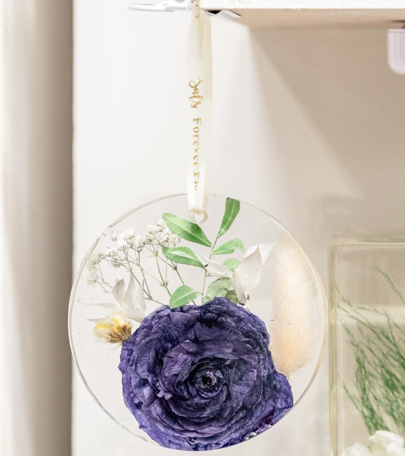 Round resin hanging ornament with preserved purple flower, greenery, and delicate white blooms, suspended on cream ribbon